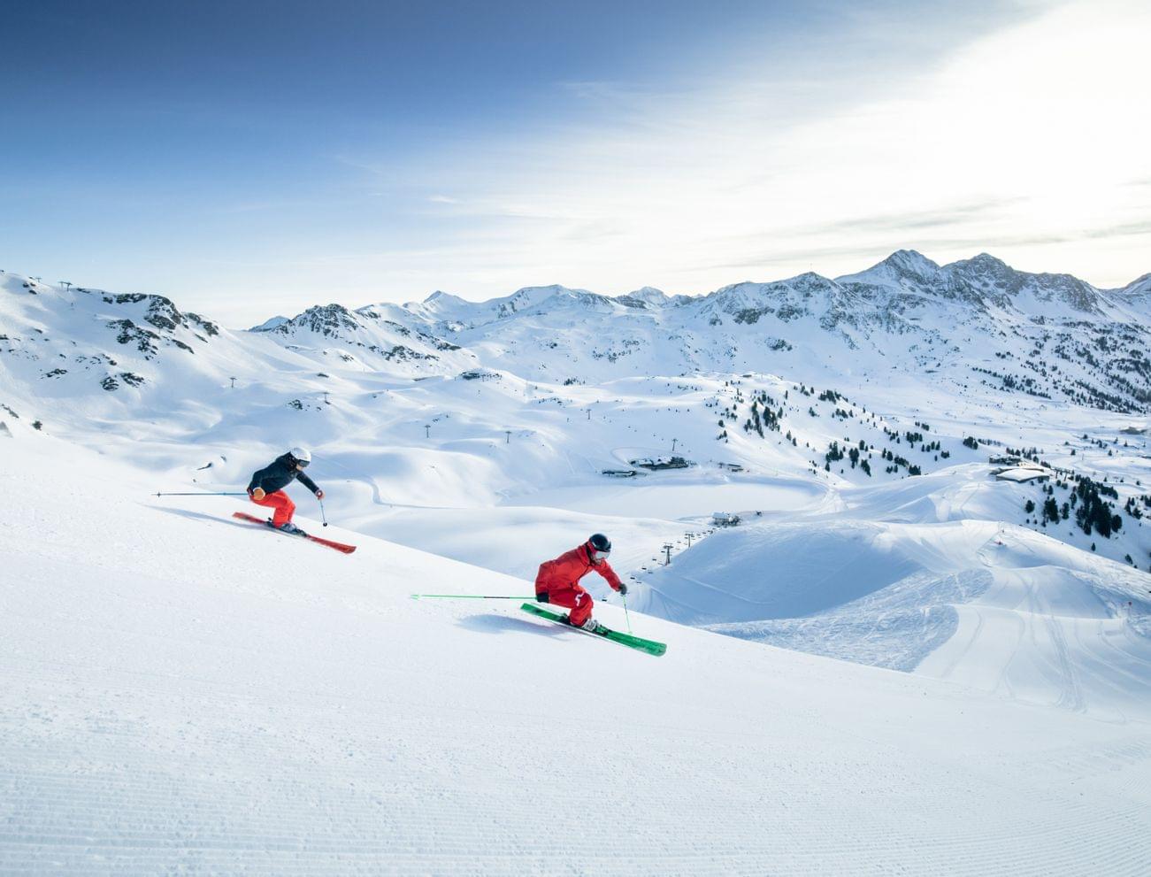 Two skiers carving down a groomed slope in a snowy alpine ski resort
