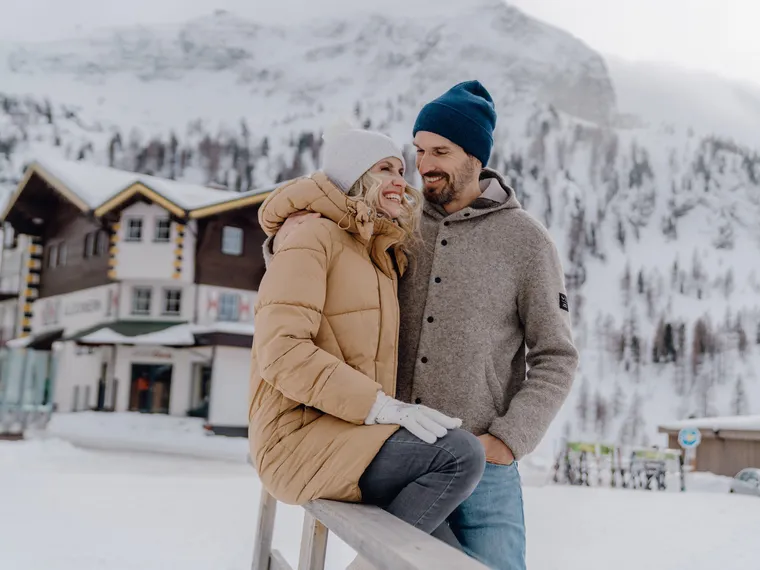 Couple in winter clothes smiling in snowy alpine village with wooden lodge