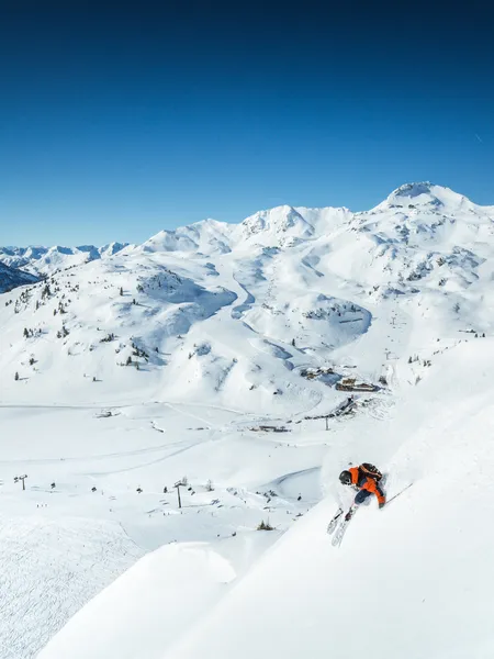 Skifahrer in orangener Jacke fährt durch frischen Pulverschnee auf steiler Alpenskipiste