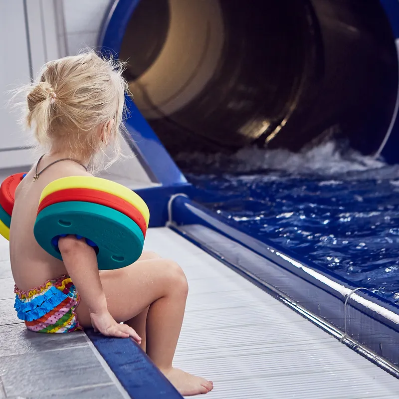 Kleines Kind mit Schwimmflügeln sitzt am Beckenrand vor einer Wasserrutsche