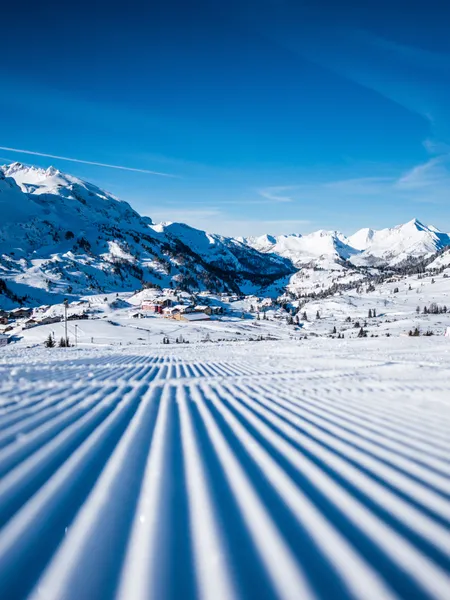 Freshly groomed ski slope with alpine village and snow-covered mountains under blue sky