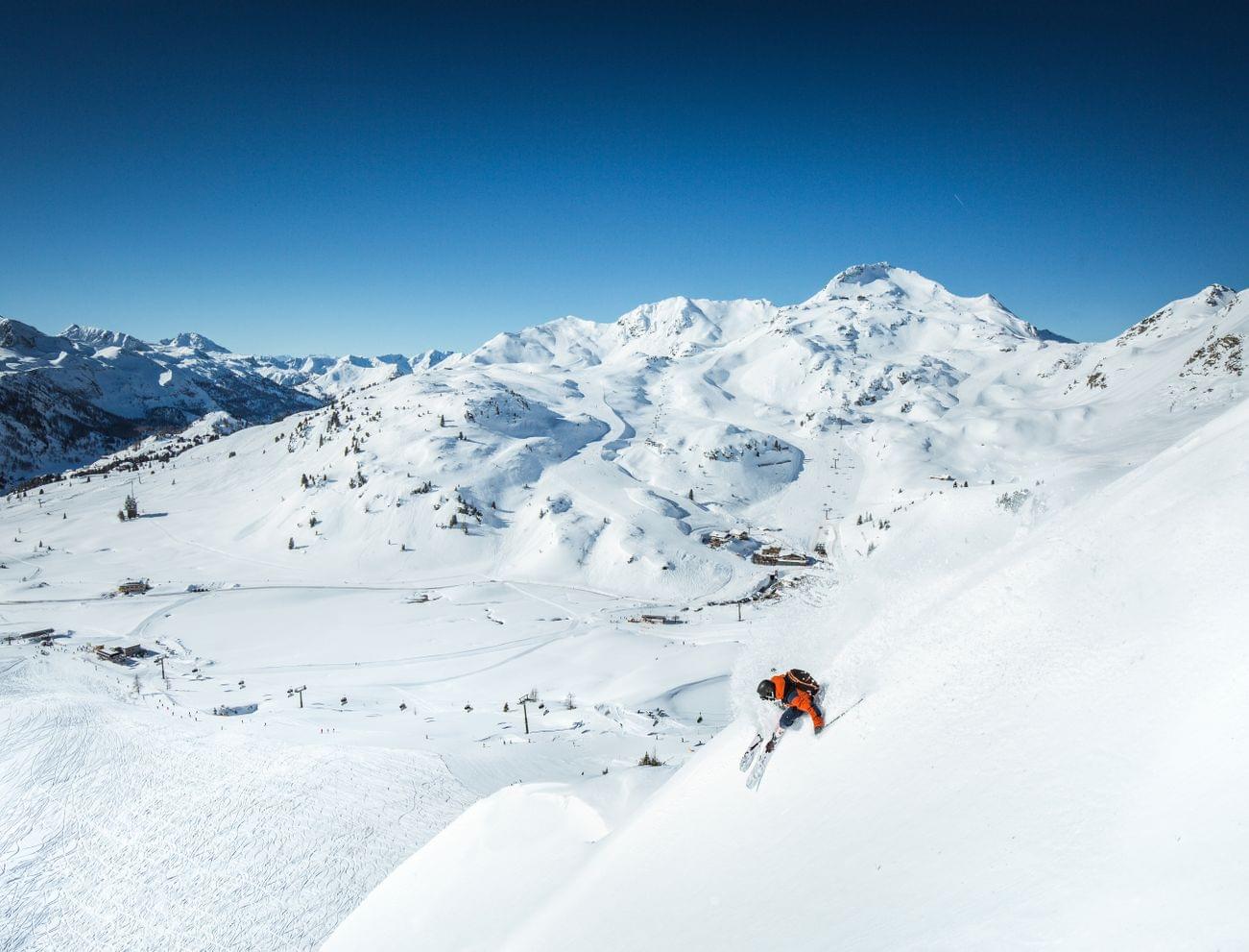 Skifahrer in orangener Jacke fährt durch frischen Pulverschnee auf steiler Alpenskipiste