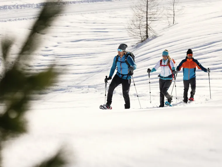 Three people snowshoe hiking through snowy mountain landscape in winter gear