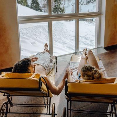 Couple relaxing on loungers indoors with panoramic snowy mountain view