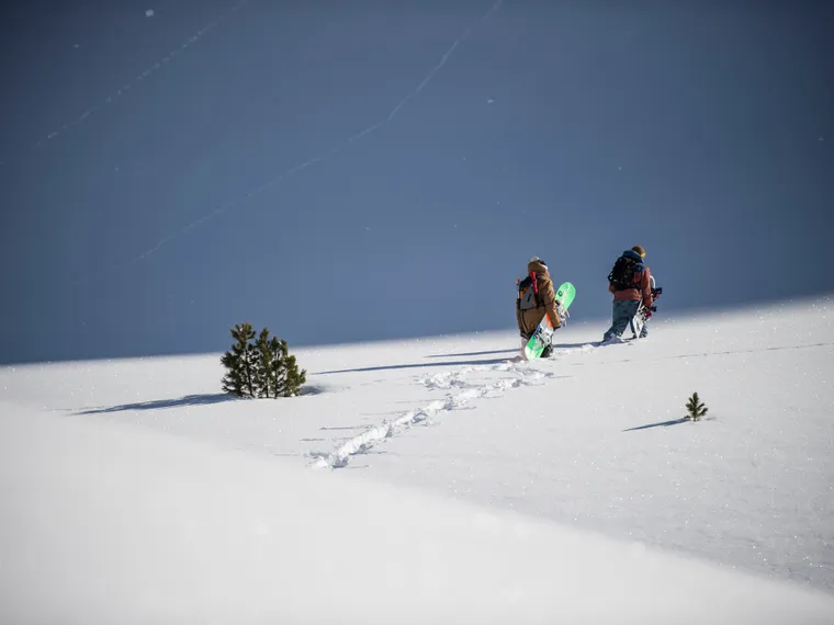 Zwei Snowboarder wandern mit Ausrüstung durch tiefen Pulverschnee am Berg