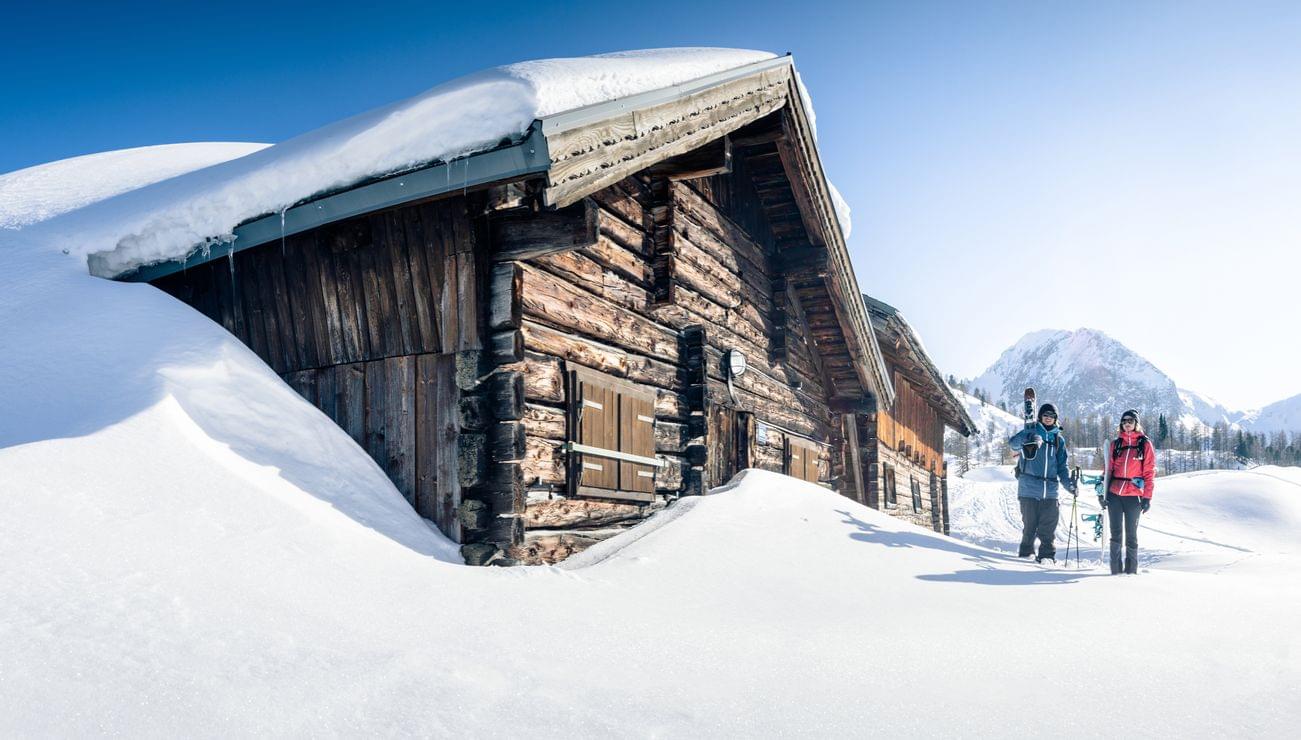 Zwei Skifahrer gehen an einer verschneiten Almhütte in den Bergen vorbei