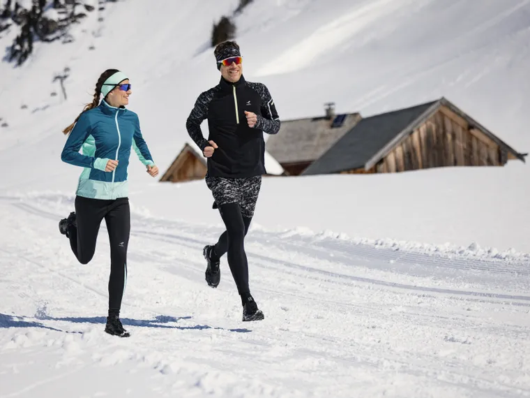 Two runners jogging on a snowy trail in bright winter conditions near alpine huts