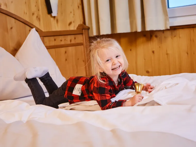 Lächelndes Kind im roten Karohemd liegt im Hotelbett und hält eine kleine goldene Glocke