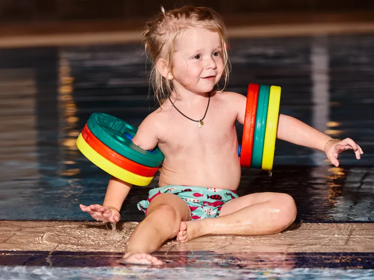 Young girl wearing colorful swim floaties sitting on poolside at indoor swimming pool