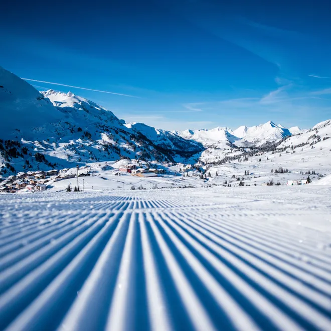 Freshly groomed ski slope with alpine village and snow-covered mountains under blue sky