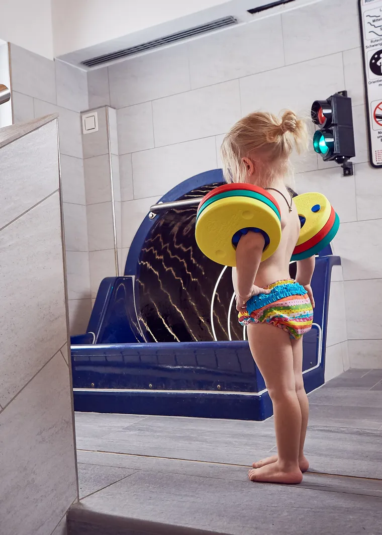 Kleines Kind mit Schwimmhilfen steht vor Rutsche im Hallenbad