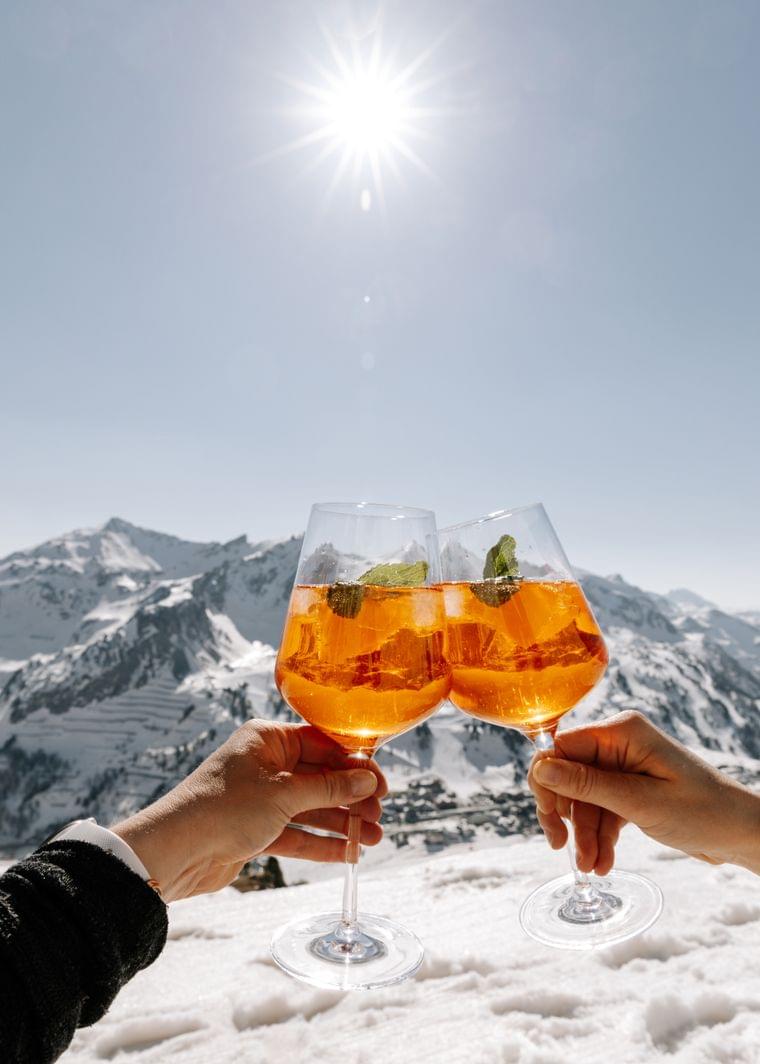Two Aperol Spritz glasses clinking in sunny snowy Alpine mountain landscape