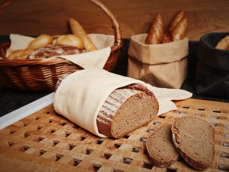 Geschnittenes Roggenbrot auf Holzbrett mit Körben voller Brötchen im Hintergrund