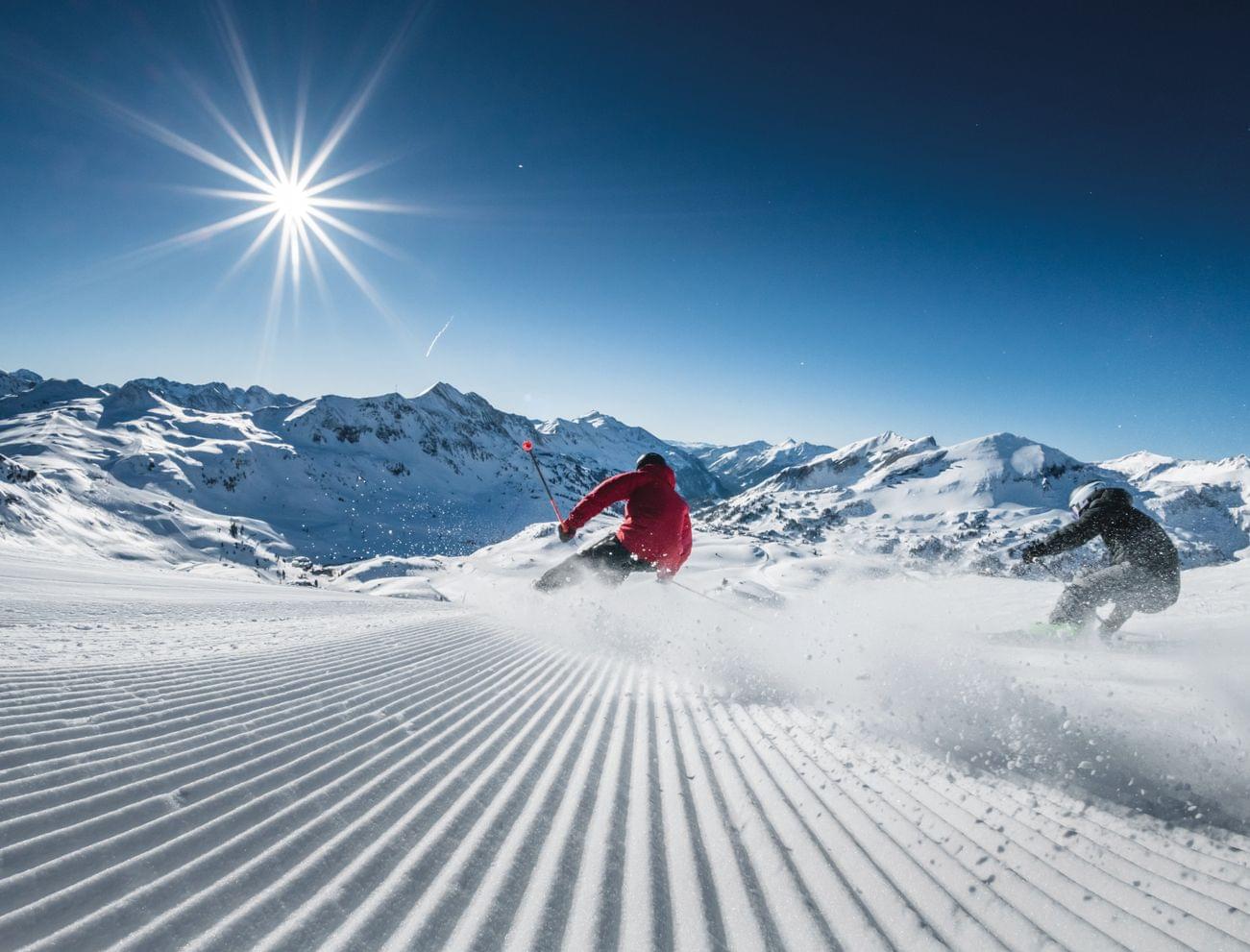 Zwei Skifahrer fahren eine präparierte Piste hinab mit schneebedeckten Bergen und greller Sonne im Hintergrund