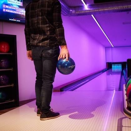 Mann mit blauer Bowlingkugel vor beleuchteter Bowlingbahn mit Neonlicht
