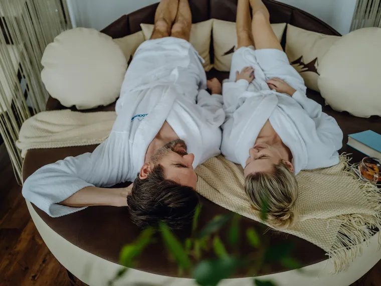 Couple in bathrobes relaxing on spa lounge bed with tea and a book