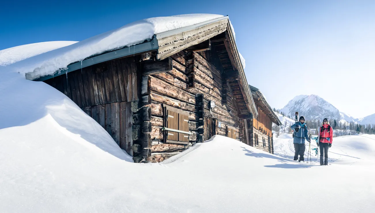 Two skiers walking past a snow-covered alpine cabin in the mountains