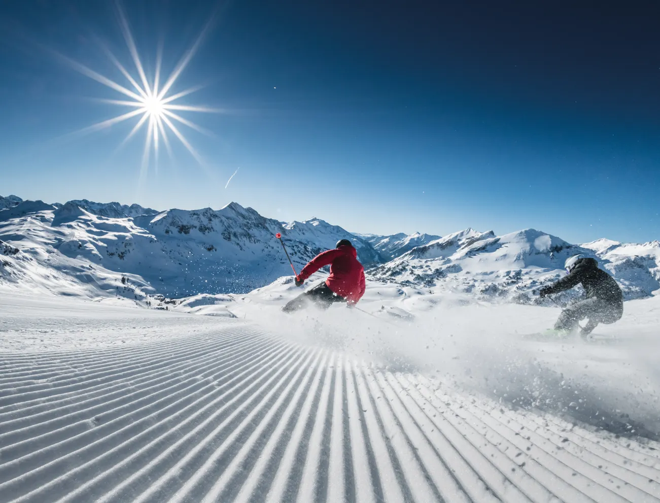 Zwei Skifahrer fahren eine präparierte Piste hinab mit schneebedeckten Bergen und greller Sonne im Hintergrund