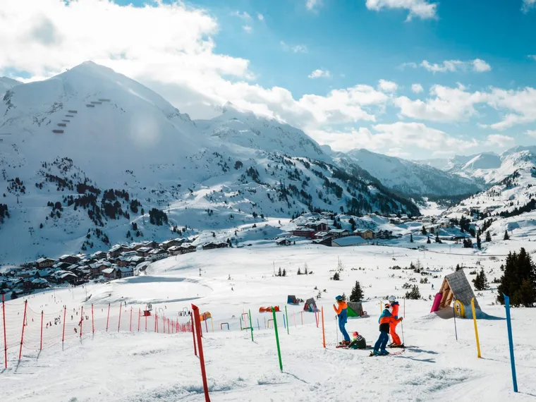 Kinder beim Skifahren an einem sonnigen Tag in einem verschneiten Alpen-Skigebiet mit Bergen im Hintergrund