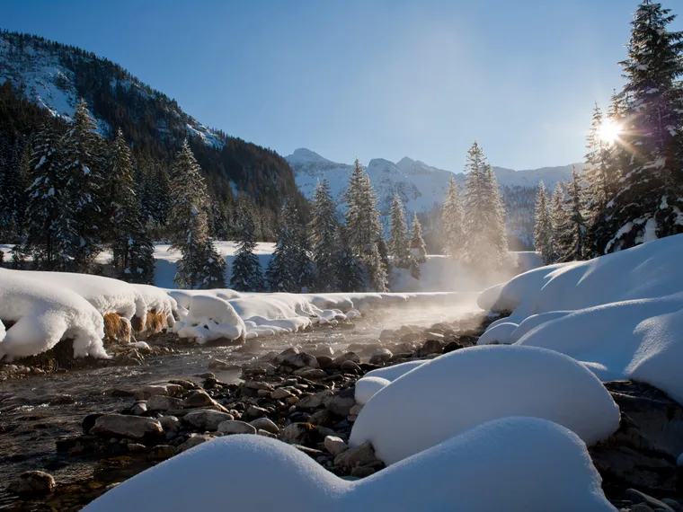 Verschneiter Alpenwald mit fließendem Fluss unter klarem blauen Himmel