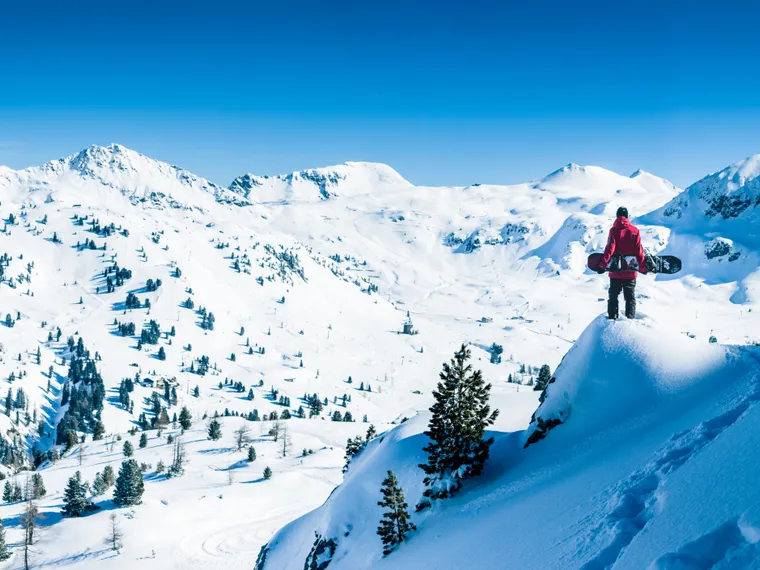 Snowboarder stands on snowy peak overlooking Alpine mountain range under clear blue sky