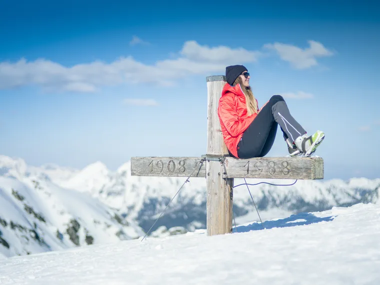 Frau im roten Anorak sitzt auf Gipfelkreuz in verschneiten Alpen