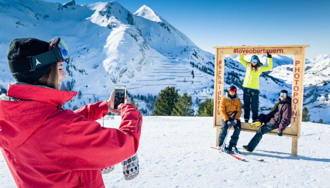 Skifahrer posieren am Obertauern-Fotopunkt in den verschneiten Alpen, während Freundin ein Foto macht