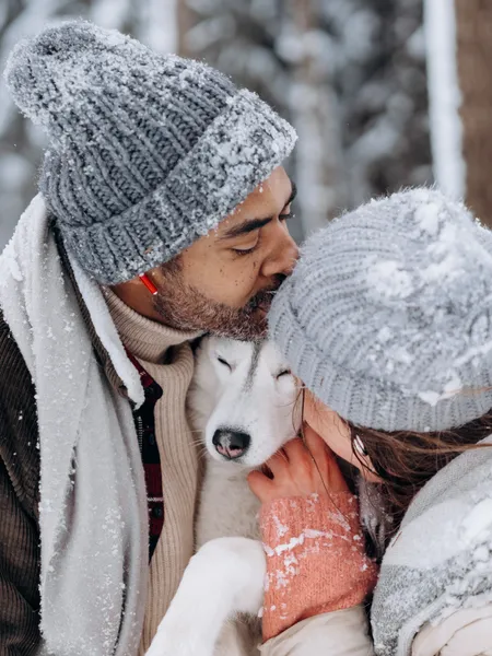 Couple in winter clothes cuddling husky dog in snowy forest