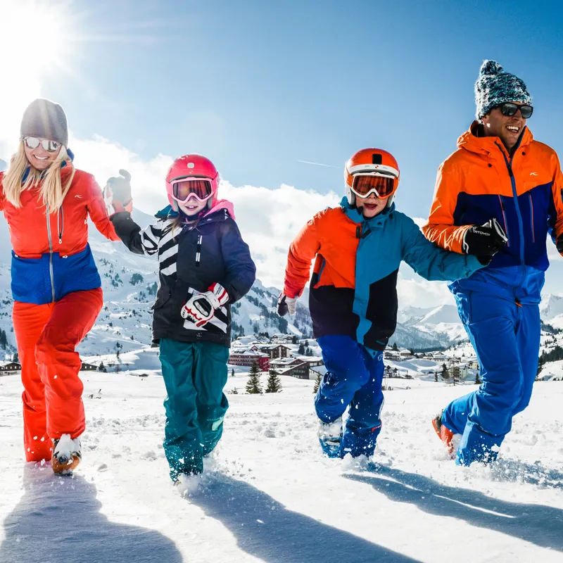 Familie in bunter Skibekleidung läuft lachend durch den Schnee in den Alpen