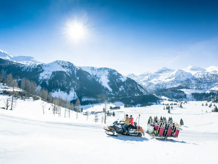 Schneemobil zieht Schlitten mit Menschen durch verschneite Alpenlandschaft bei strahlender Sonne