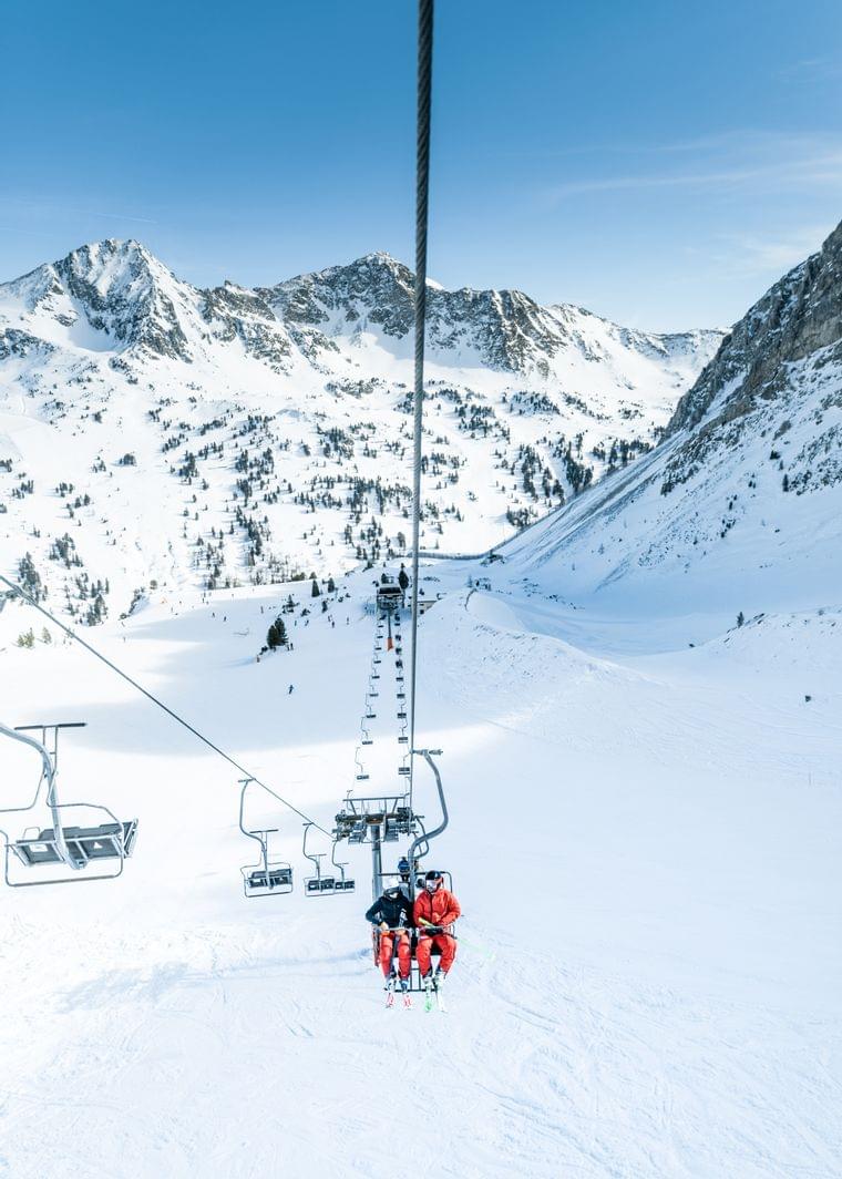Skiers on a mountain chairlift with snowy alpine peaks in the background