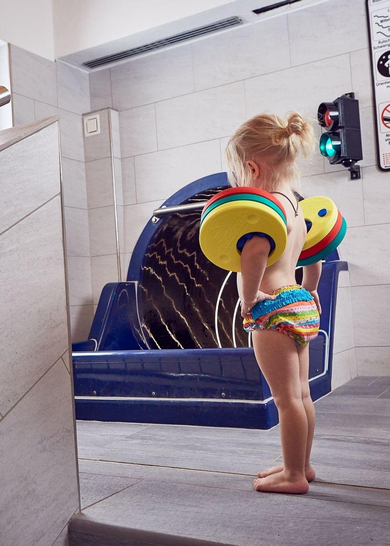 Small child with floatation armbands stands near indoor pool slide
