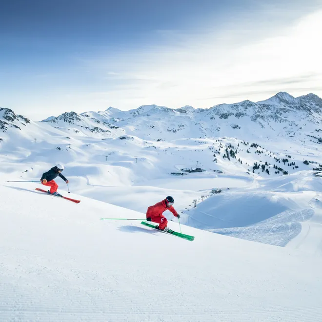 Two skiers carving down a groomed slope in a scenic alpine ski resort
