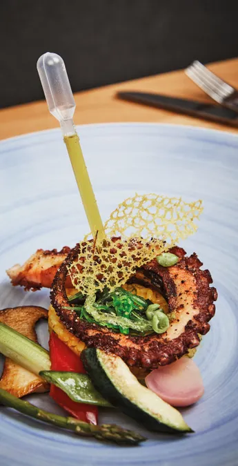 Gourmet grilled octopus dish with vegetables, seaweed, rice, and decorative lace crisp on a blue plate