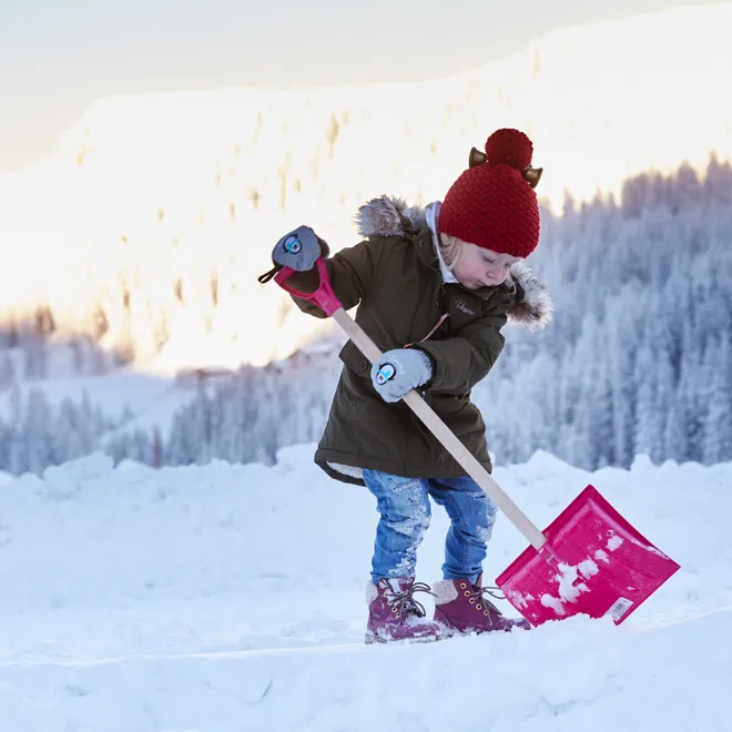 Kind mit roter Mütze schaufelt Schnee in winterlicher Berglandschaft