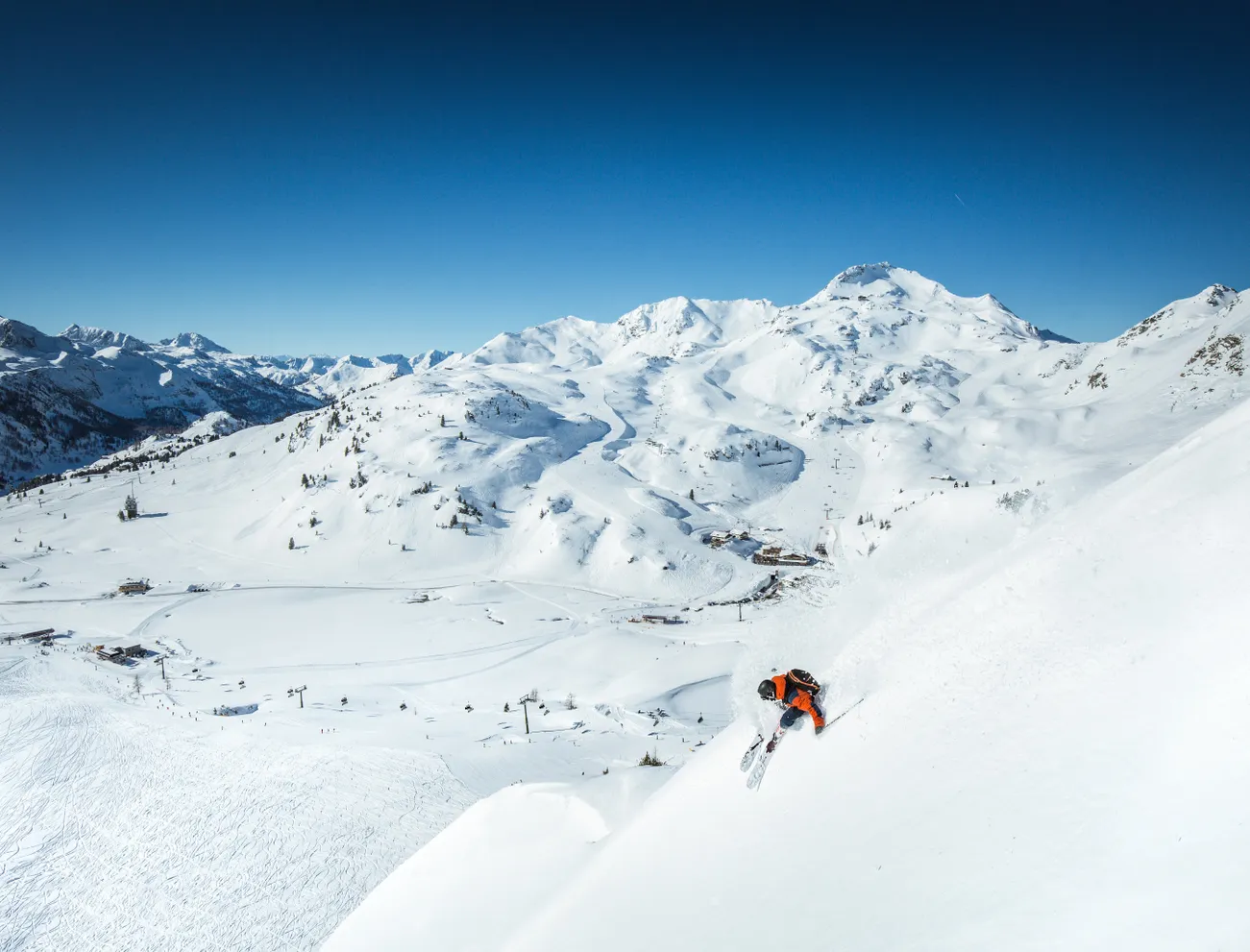 Skifahrer in orangener Jacke fährt durch frischen Pulverschnee auf steiler Alpenskipiste