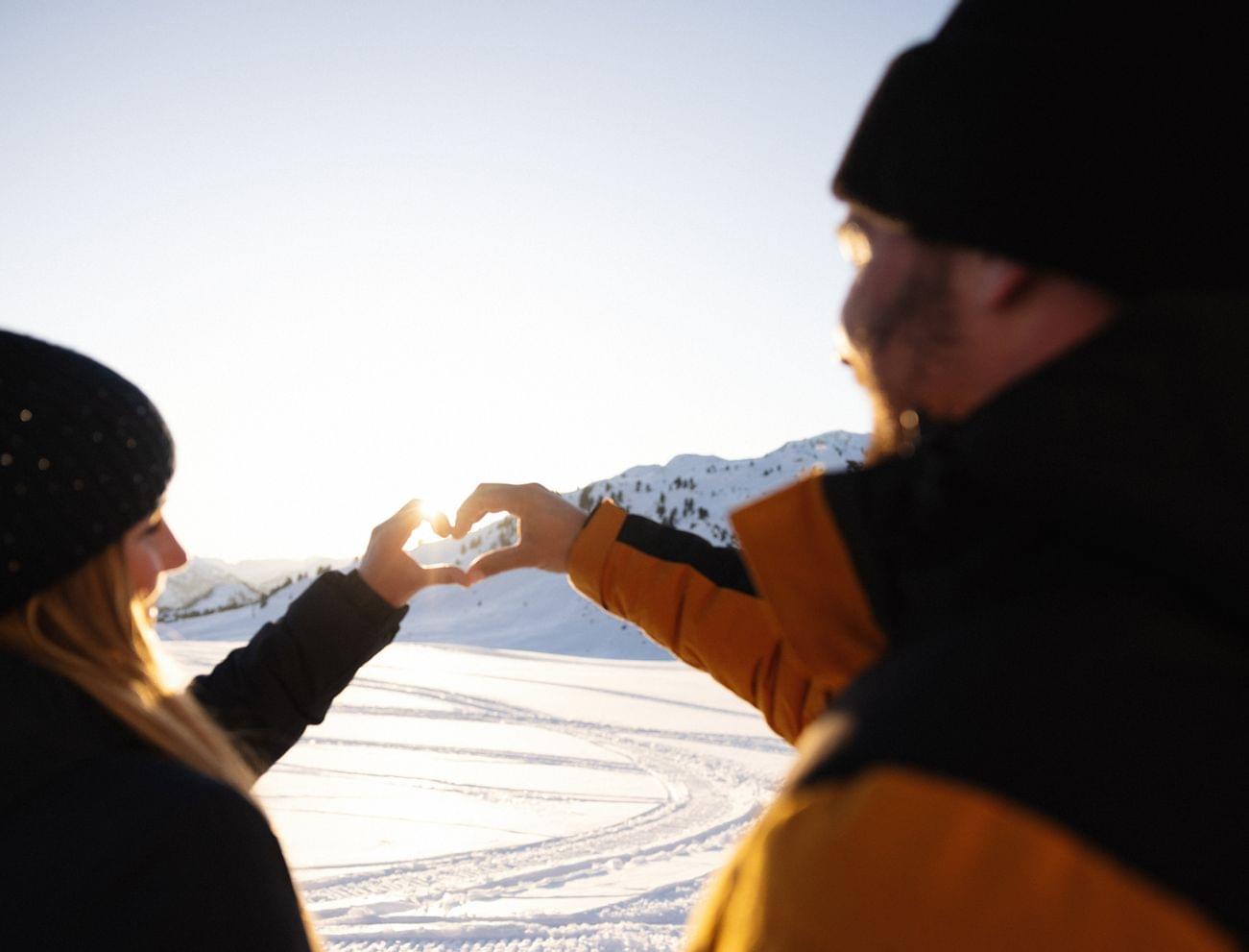 Couple making heart shape with hands against snowy mountain sunset