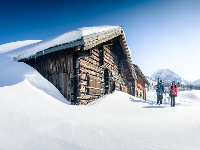 Two skiers walking past a snow-covered alpine cabin in the mountains